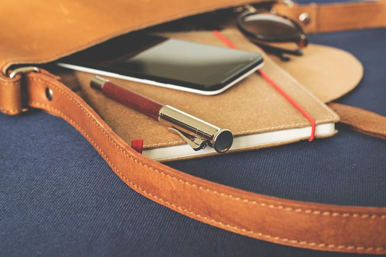 Black smartphone with its screen turned off resting on a notebook beside a fountain pen in a tan leather bag.