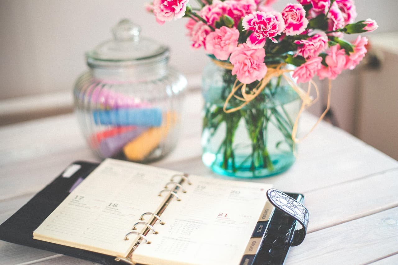 Open paper planner beside pink flowers and a glass jar on a white desk, representing bills and dates to consider before payday.